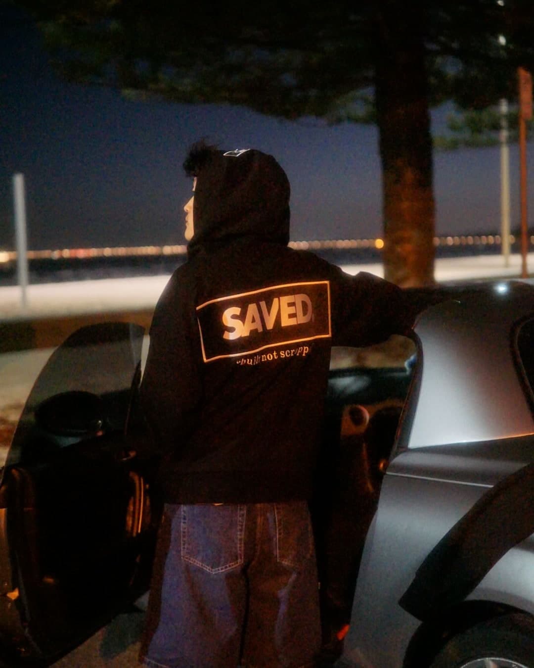 A SAVED. hoodie worn at the waterfront at night, the reflective back print lit up beside an open car door