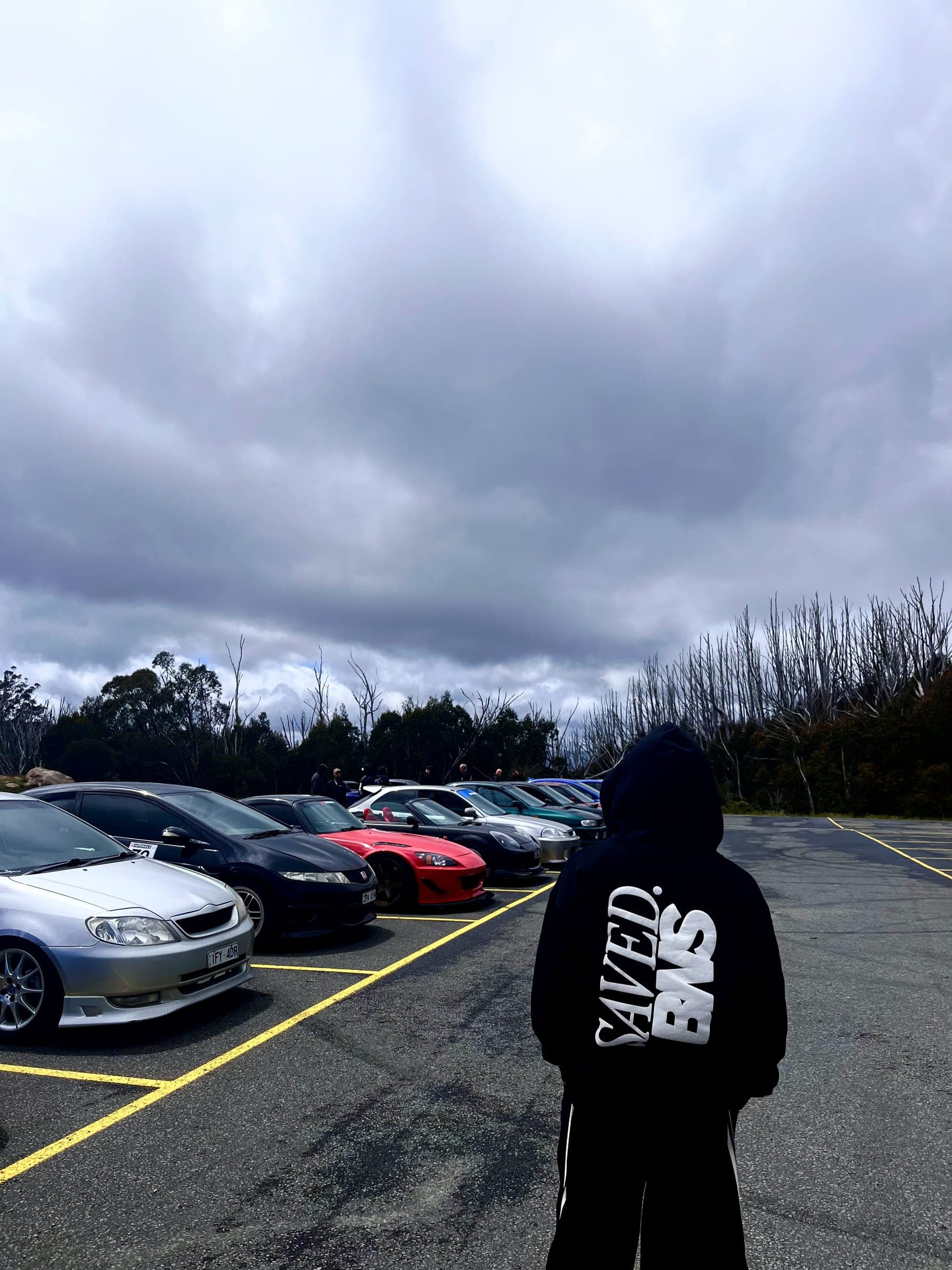 A SAVED. BNS hoodie worn at a car meet, a row of JDM cars parked under heavy cloud
